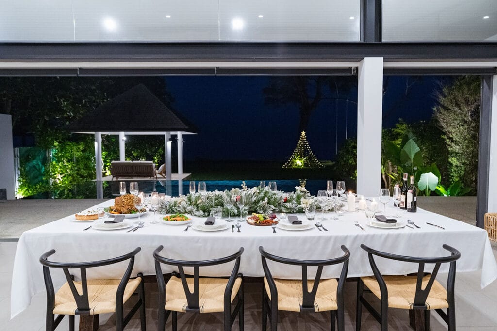Alt: Nighttime beachfront Christmas dinner at Twin Villas Natai, with a long holiday table overlooking the pool, garden, and illuminated outdoor Christmas tree for a serene Christmas villa Thailand celebration.
