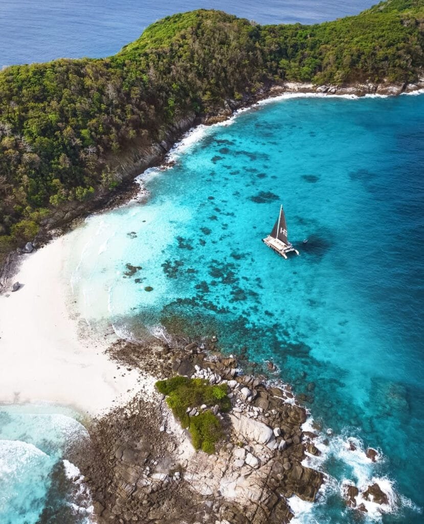 Aerial view of a luxury sailing catamaran Hype Boat anchored in a pristine turquoise bay with a white sand beach, perfect for a private family day trip.