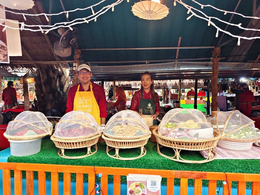  Local vendors at Long Lae Sunday Market presenting traditional Thai street food snacks in woven baskets covered with protective netting.
