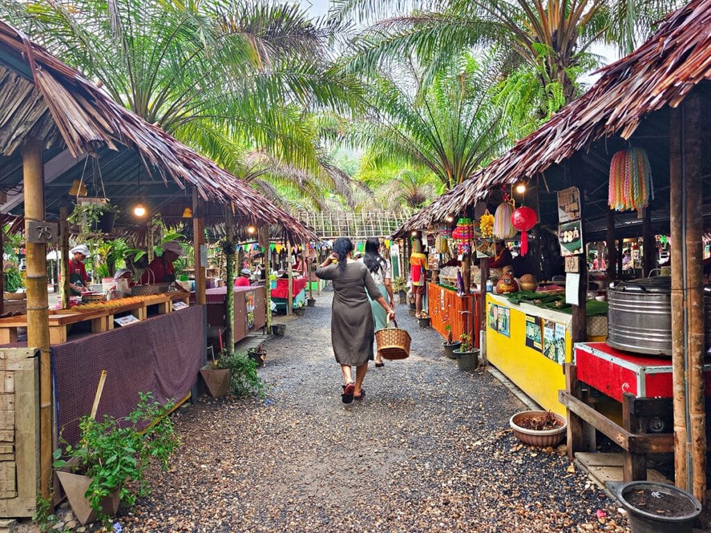 A vibrant walking path at Long Lae Sunday Market in Kap Pong, showing traditional thatched-roof stalls and local visitors.
