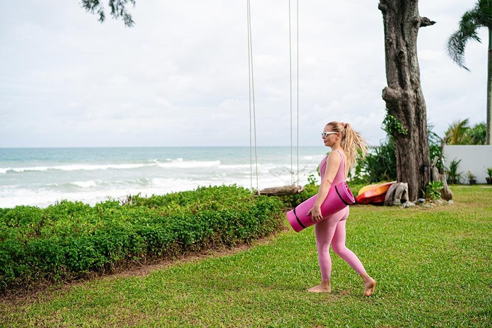 Retreat Villas Near Phuket – A Private Beachfront Wellness Sanctuary at Twin Villas Natai | Twin Villas Natai Retreat participant carrying a yoga mat across a beachfront garden before a wellness session.